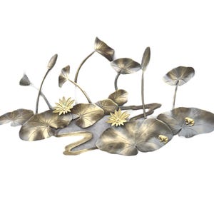 Puede incluir: Una escultura de metal de una escena de jardín acuático con hojas de nenúfar, flores de loto y pequeñas ranas. La escultura presenta tonos dorados y plateados, con diseños detallados de hojas y flores. El tema es la naturaleza y la tranquilidad.