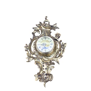 May include: Ornate brass wall clock with a round face featuring Roman numerals. The clock is surrounded by intricate floral and scrollwork designs. The clock's hands are blue, and the face has a light-colored background.