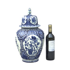 Puede incluir: Un jarrón de cerámica grande, azul y blanco con un diseño floral y una pequeña figura de león en la tapa. Una botella de vino tinto está al lado del jarrón.