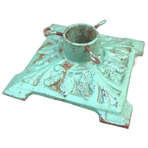 May include: A distressed, square Christmas tree stand with a turquoise finish and ornate leaf detailing. The stand features a central cylinder to hold the tree trunk and has rusty accents.
