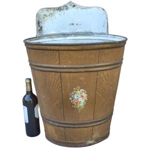 May include: An antique wooden wash tub with a light blue metal backsplash. The tub has a floral design and is next to a dark wine bottle. The wood has a natural grain and is held together with metal bands.