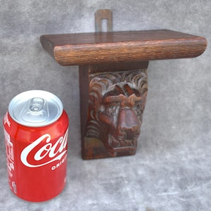 May include: A small, dark brown wooden shelf with a carved lion's head supporting the shelf. The shelf is rectangular and has a metal bracket for hanging. A red Coca-Cola can is next to the shelf.