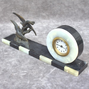 May include: An Art Deco clock with a black and white marble base and a round clock face. A bronze sculpture of two birds in flight is on the left side. The clock face has gold-colored trim and black numerals.
