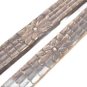 May include: Close-up of two decorative wooden trim pieces. Each piece features a carved floral design in the centre and a textured, scale-like pattern along the edges. The trim has a weathered, antique appearance with brown and grey tones.