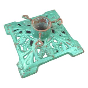 Op de afbeelding: Een vintage kerstboomstandaard van gietijzer, geschilderd in een verweerd zeegroen met rode accenten. De basis heeft een bladeren ontwerp. De centrale metalen beker houdt de boomstam vast, met drie metalen lussen.
