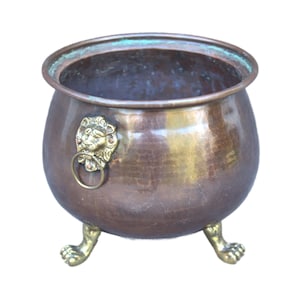 Puede incluir: Un caldero de cobre martillado con una cabeza de león de bronce y un asa de anillo. El caldero tiene tres patas de león de bronce. El interior tiene una pátina oscura y el exterior está pulido.