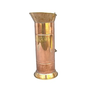 May include: A copper and brass cylindrical container with a flared top and a rounded base. The word "KAN" is embossed on the side. The container has a small handle and is likely an antique or vintage item.