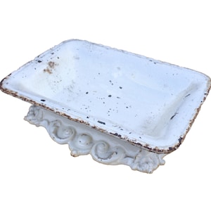 May include: A rectangular white enameled metal soap dish with a raised, ornate base featuring scrollwork details. The edges of the dish show visible brown rust and wear, with small dark speckles scattered across the interior surface.