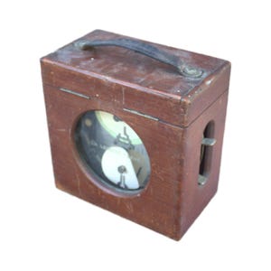 May include: A vintage wooden box with a round gauge and a leather handle. The box is rectangular with a dark brown finish and a metal clasp on the side. The gauge has a white face with black markings.