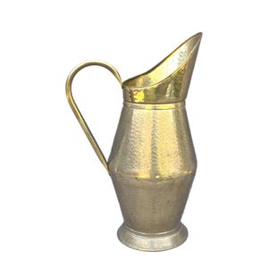 May include: A vintage metal pitcher with a hammered texture. The pitcher has a curved handle and a spout with a gold-colored rim. The body of the pitcher is a silver color, and it sits on a round base. The pitcher is likely made of brass or a similar metal.