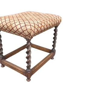May include: A square stool featuring a cushioned seat upholstered in a beige fabric with a dark red diamond grid pattern. The seat is trimmed with small metal nailheads. The base consists of four dark brown wooden legs with a twisted design.