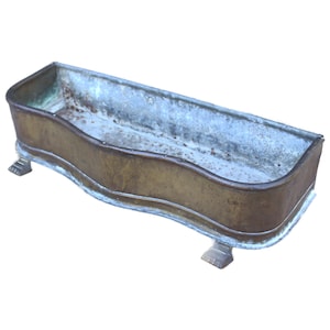 Puede incluir: Una jardinera rectangular de latón antiguo con un diseño suavemente curvado. La jardinera se apoya en cuatro pequeños pies decorativos. El interior muestra signos de edad y uso, con un acabado patinado.