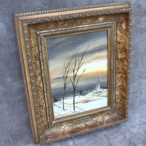 Puede incluir: Pintura de paisaje enmarcada que representa una escena invernal con árboles desnudos y un campo nevado bajo un cielo nublado. La pintura está en un marco dorado ornamentado.