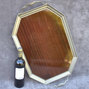 Op de afbeelding: Een achthoekige serveerschaal met een donker houten nerfcentrum en een goudkleurig metalen frame. De schaal heeft decoratieve handgrepen en staat naast een fles rode wijn.