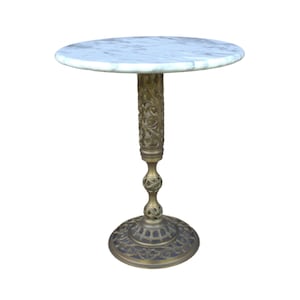 Puede incluir: Una pequeña mesa auxiliar redonda con tapa de mármol blanco. La base ornamentada es de metal color latón con intrincados diseños florales y de volutas. La mesa mide aproximadamente 71 cm de altura.