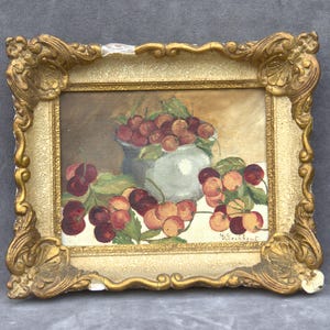 Puede incluir: Una pintura antigua de naturaleza muerta de cerezas en un cuenco blanco y sobre una mesa. La pintura está en un marco dorado ornamentado con algunos daños. Las cerezas son rojas y amarillas con hojas verdes.