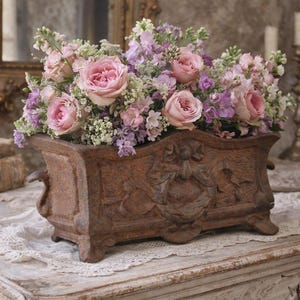 Könnte beinhalten: Ein rechteckiger, verwitterter brauner Pflanzkübel aus Gusseisen mit einem verzierten Reliefdesign aus einem zentralen Kranz und einer Schleife. Der Kübel ist mit hellrosa Rosen, violetten Levkojen und kleinen weißen Blüten gefüllt.