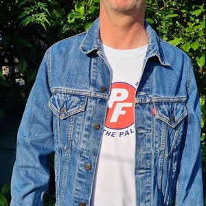 Può includere: Una giacca di jeans blu con una t-shirt bianca sotto. La t-shirt ha un cerchio rosso con le lettere "PF" in bianco e il testo "The Pal" sotto.