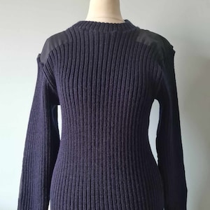 Może przedstawiać: Granatowy sweter z prążkowanego dzianiny z czarnymi naszywkami na ramionach. Sweter ma okrągły dekolt i długie rękawy.