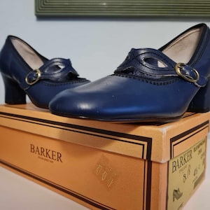 Può includere: Un paio di scarpe in pelle blu navy con chiusura a fibbia. Le scarpe sono appoggiate su una scatola marrone con la scritta "Barker" stampata su di essa.