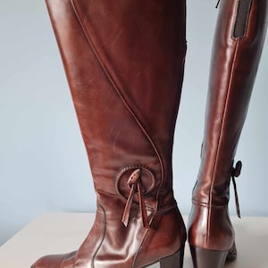May include: A pair of brown leather knee-high boots with a block heel and a decorative tie detail on the front of one boot.