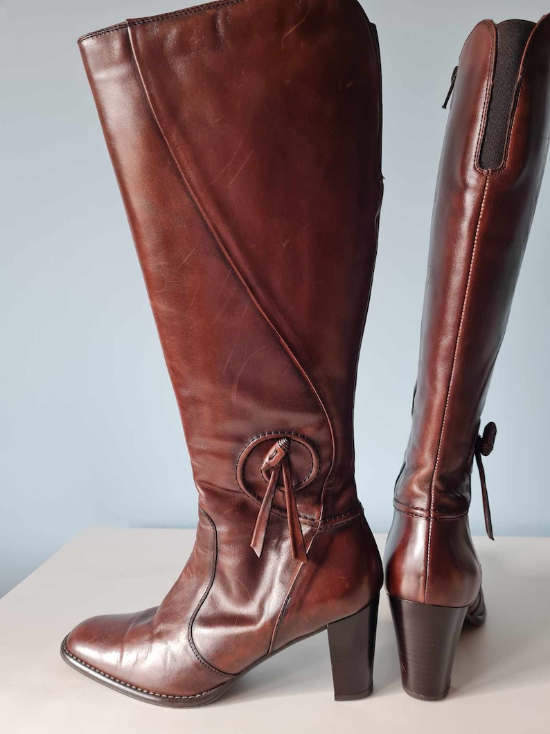 May include: A pair of brown leather knee-high boots with a block heel and a decorative tie detail on the front of one boot.