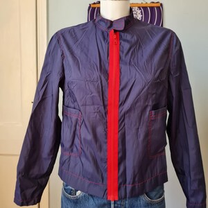 May include: A vintage navy blue jacket with a bright red zipper and matching red stitching. The jacket has two front pockets and a stand-up collar. The jacket is displayed on a mannequin with blue jeans.