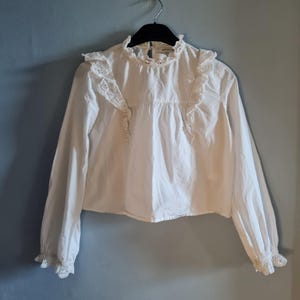 May include: A white long-sleeve blouse with a high, ruffled collar and lace detailing. The blouse has a button-down front and ruffled accents on the shoulders and cuffs. The garment is hanging on a black hanger against a blue wall.