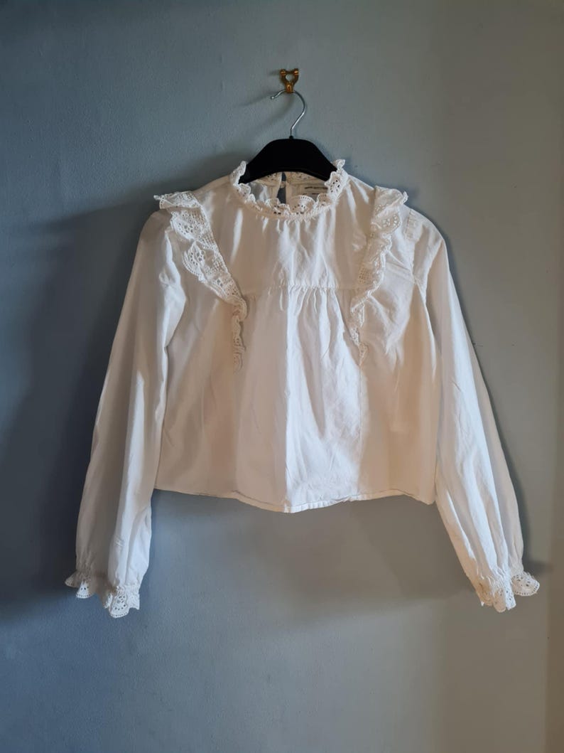 May include: A white long-sleeve blouse with a high, ruffled collar and lace detailing. The blouse has a button-down front and ruffled accents on the shoulders and cuffs. The garment is hanging on a black hanger against a blue wall.