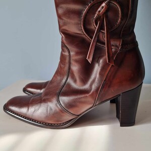 May include: Brown leather knee-high boots with a chunky heel and a decorative strap detail at the top of the boot.