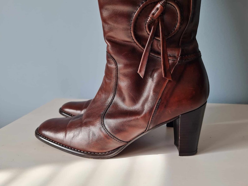 May include: Brown leather knee-high boots with a chunky heel and a decorative strap detail at the top of the boot.