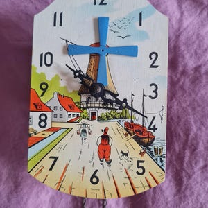 May include: A wall clock with a light-coloured wooden face featuring a printed scene of a windmill, houses, and a person walking. The clock has black hour and minute hands, a blue windmill-shaped second hand, and black numbers from 1 to 12.