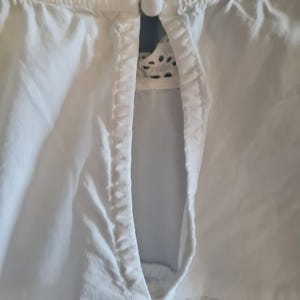 May include: Close-up of a white blouse with a keyhole neckline. The blouse features a delicate lace trim around the collar and a small, round button closure. The fabric appears to be a lightweight cotton or linen.