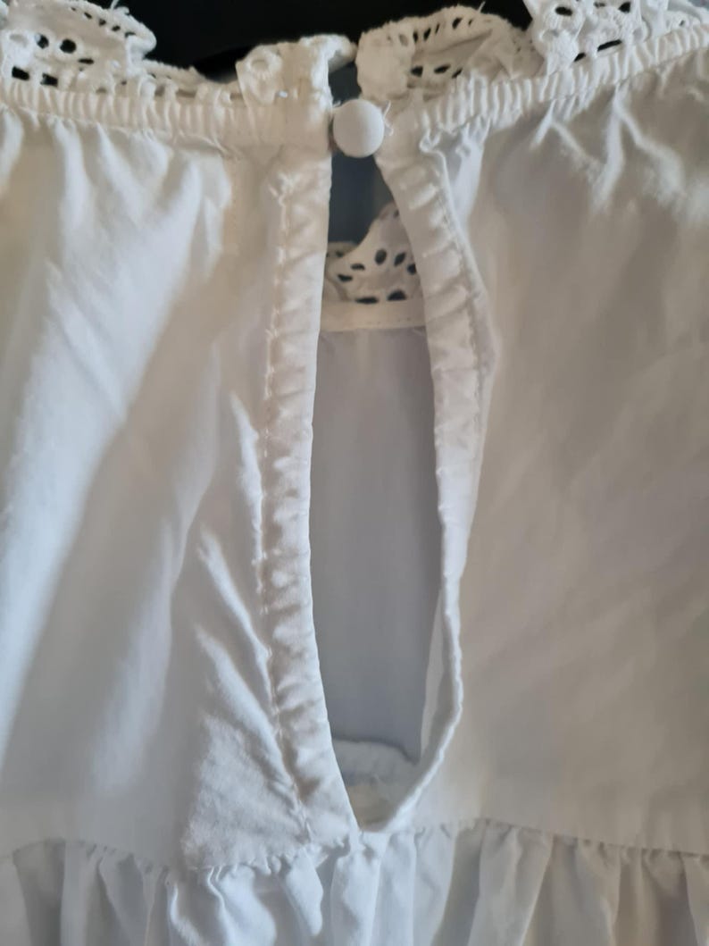 May include: Close-up of a white blouse with a keyhole neckline. The blouse features a delicate lace trim around the collar and a small, round button closure. The fabric appears to be a lightweight cotton or linen.