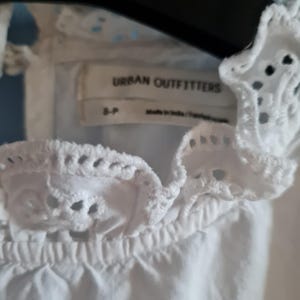 May include: Close-up of a white eyelet top with a ruffled neckline. The top features the brand name "URBAN OUTFITTERS" on a tag. The top is made in India.