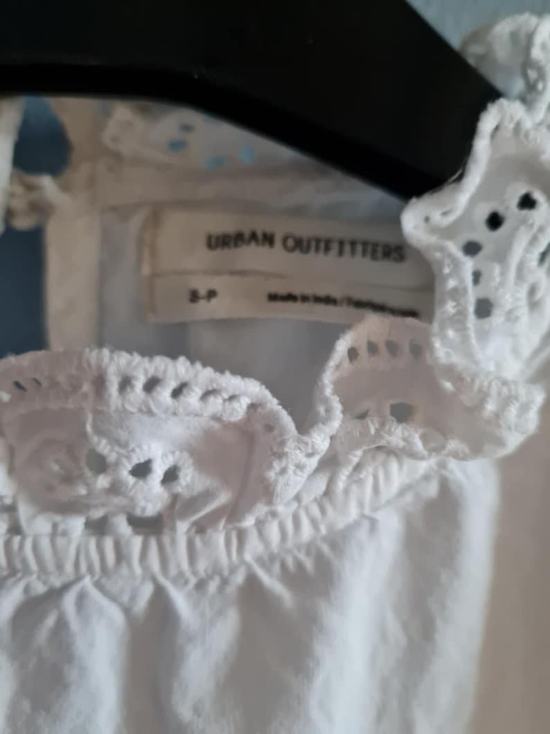 May include: Close-up of a white eyelet top with a ruffled neckline. The top features the brand name "URBAN OUTFITTERS" on a tag. The top is made in India.
