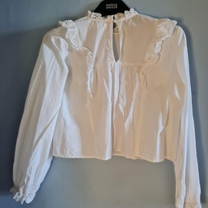 May include: White long-sleeve blouse with a high neckline and delicate lace trim. The blouse features a keyhole back and is hanging on a black hanger. The sleeves have lace detailing at the cuffs.
