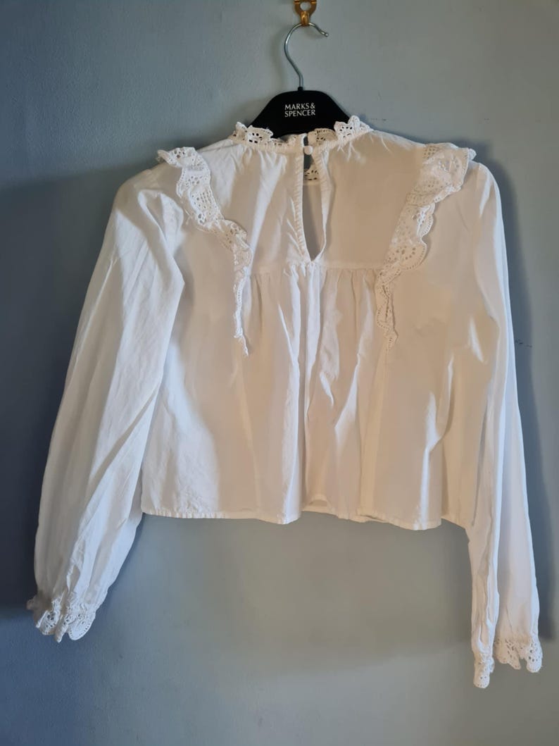 May include: White long-sleeve blouse with a high neckline and delicate lace trim. The blouse features a keyhole back and is hanging on a black hanger. The sleeves have lace detailing at the cuffs.