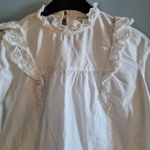 May include: White long-sleeve blouse with a high, ruffled collar and lace detailing on the shoulders and upper chest. The blouse has a gathered bodice and a simple, elegant design, suitable for various occasions.