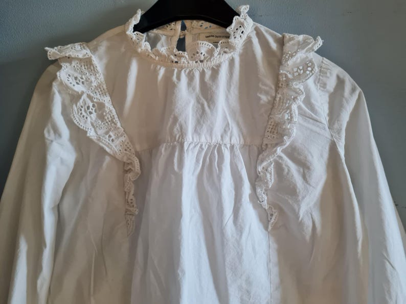 May include: White long-sleeve blouse with a high, ruffled collar and lace detailing on the shoulders and upper chest. The blouse has a gathered bodice and a simple, elegant design, suitable for various occasions.