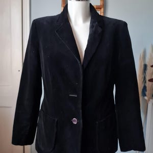 Op de afbeelding: Een zwarte fluwelen blazer met een revers, twee voorzakken en een sluiting met één knoop. De blazer wordt getoond op een mannequin, die de getailleerde pasvorm en het klassieke ontwerp laat zien. De jas is een veelzijdig kledingstuk.