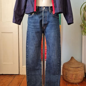 Könnte beinhalten: Eine mittelblaue Jeans aus Denim im klassischen Five-Pocket-Stil mit Knopfleiste und geradem Bein, kombiniert mit einer marineblauen Windjacke, die einen leuchtend roten Reißverschluss in der vorderen Mitte aufweist.