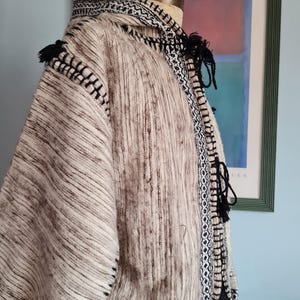 May include: A hooded cloak featuring a textured beige and brown marled knit pattern. The garment is accented with black and white geometric trim along the hood, front opening, and hem, and includes black tassel ties at the neck and shoulders.