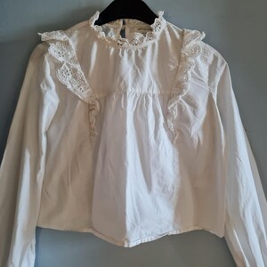 May include: White long-sleeved blouse with a high, ruffled collar and lace detailing. The blouse features lace trim on the shoulders and a gathered bodice. The garment is hanging on a black hanger against a blue-gray wall.