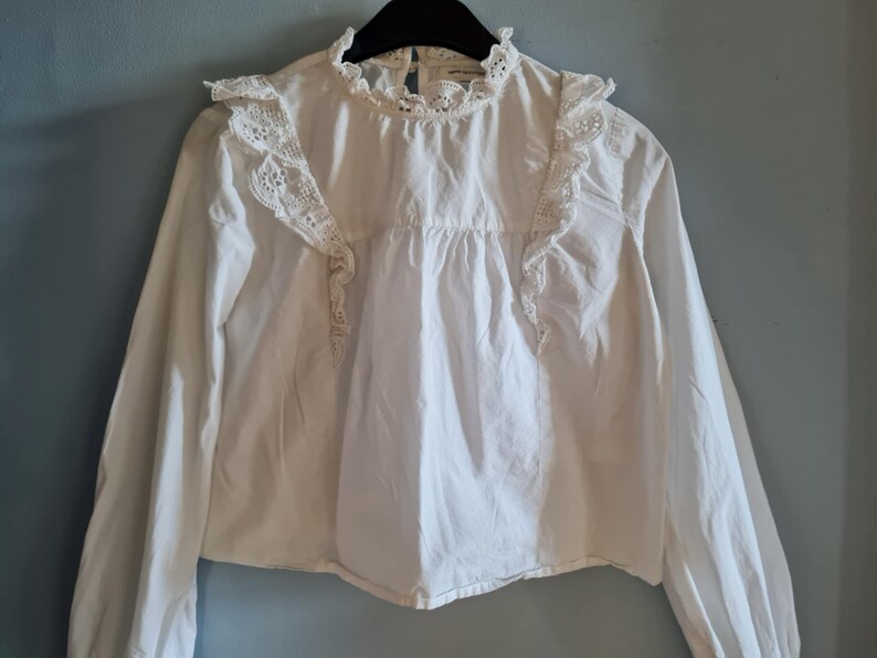 May include: White long-sleeved blouse with a high, ruffled collar and lace detailing. The blouse features lace trim on the shoulders and a gathered bodice. The garment is hanging on a black hanger against a blue-gray wall.