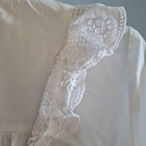 May include: Close-up of a white blouse with a detailed, eyelet lace trim along the shoulder and sleeve. The blouse has a ruffled edge and a delicate, textured appearance. The fabric appears to be lightweight and breathable.
