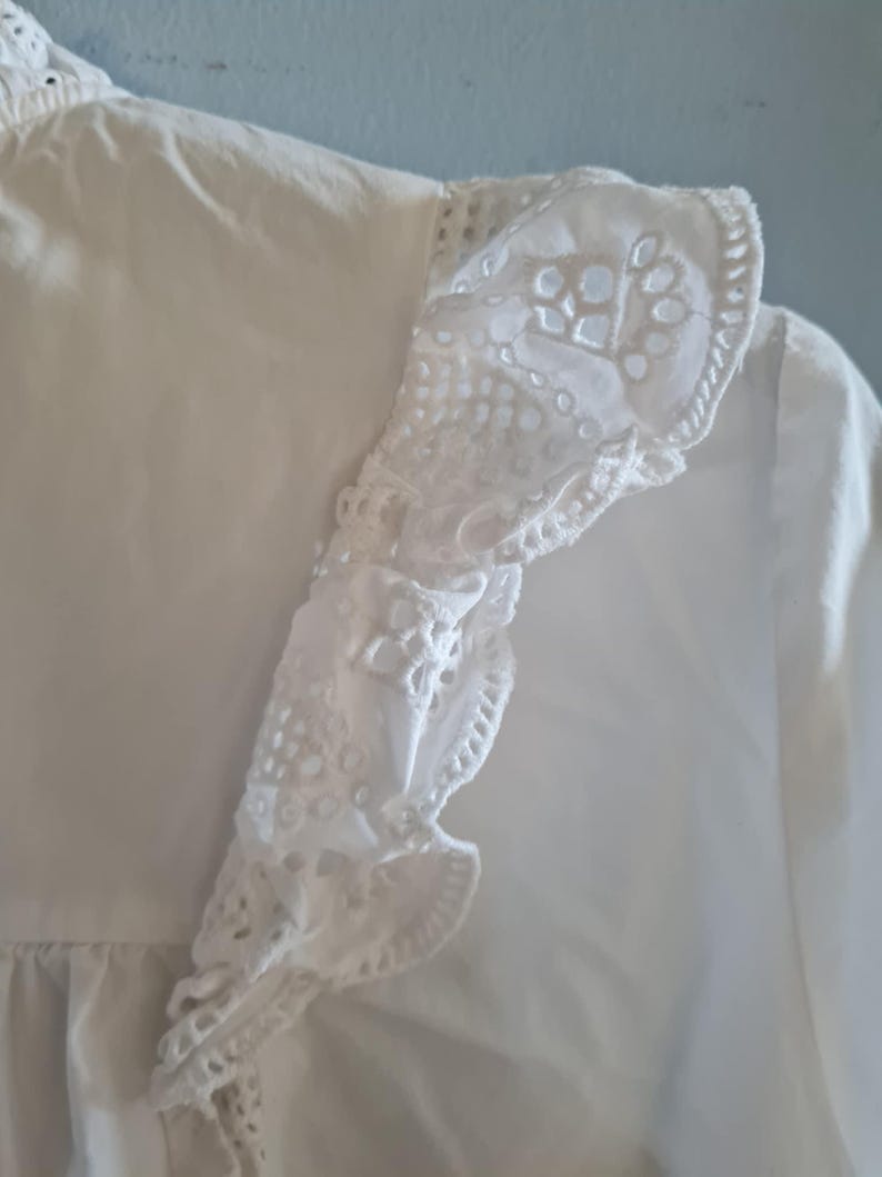 May include: Close-up of a white blouse with a detailed, eyelet lace trim along the shoulder and sleeve. The blouse has a ruffled edge and a delicate, textured appearance. The fabric appears to be lightweight and breathable.
