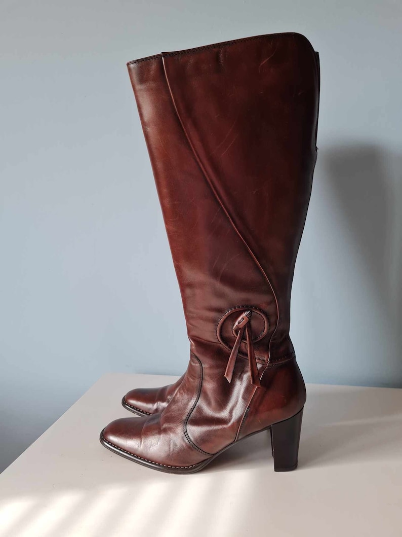 May include: Brown leather knee-high boots with a block heel and a decorative tie detail at the top of the shaft.