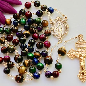 May include: A rosary with multi-colored tiger eye beads, gold-toned chain, and a gold crucifix. The beads are in shades of red, green, blue, and brown. The centerpiece features an image of the Virgin Mary.