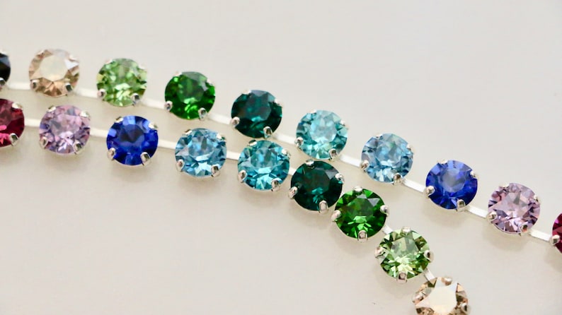 Puede incluir: Primer plano de una cadena plateada con una variedad de piedras de cristal redondas y coloridas. Las piedras de cristal est&aacute;n dispuestas en un patr&oacute;n de degradado, comenzando con morado y terminando con verde.